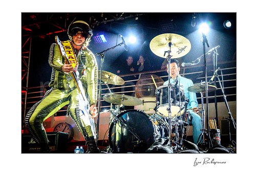 Horizontal color image of Stig Pedersen and Laust Sonne performing live on stage, with Pedersen in a reflective space suit holding his bass and Sonne playing drums under bright concert lights