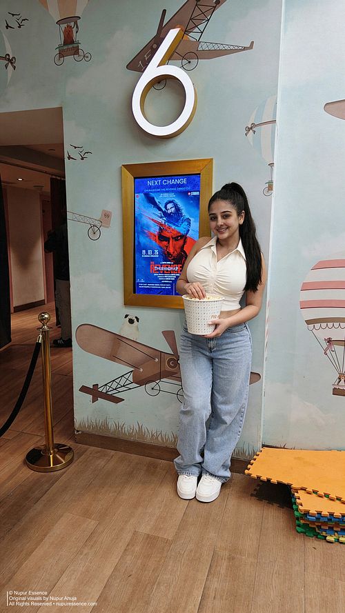 Nupur Ahuja posing with a popcorn bucket in a cinema lobby wearing a white halter top and light wash denim jeans, published by Nupur Essence