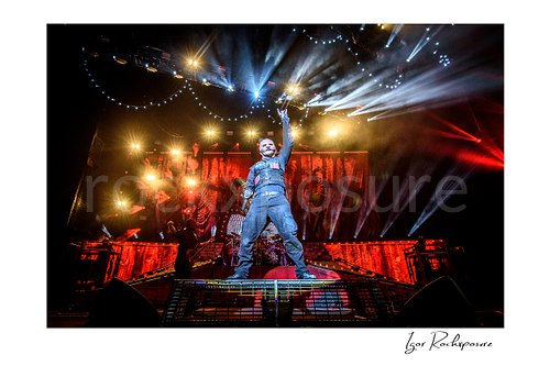 Horizontal color image of Corey Taylor of Slipknot standing center stage with one arm raised beneath dramatic white and red concert lights