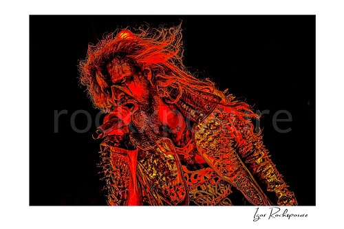 Horizontal color image of Rob Zombie performing live on stage under intense red lighting with wild hair, a studded jacket, and a handheld microphone