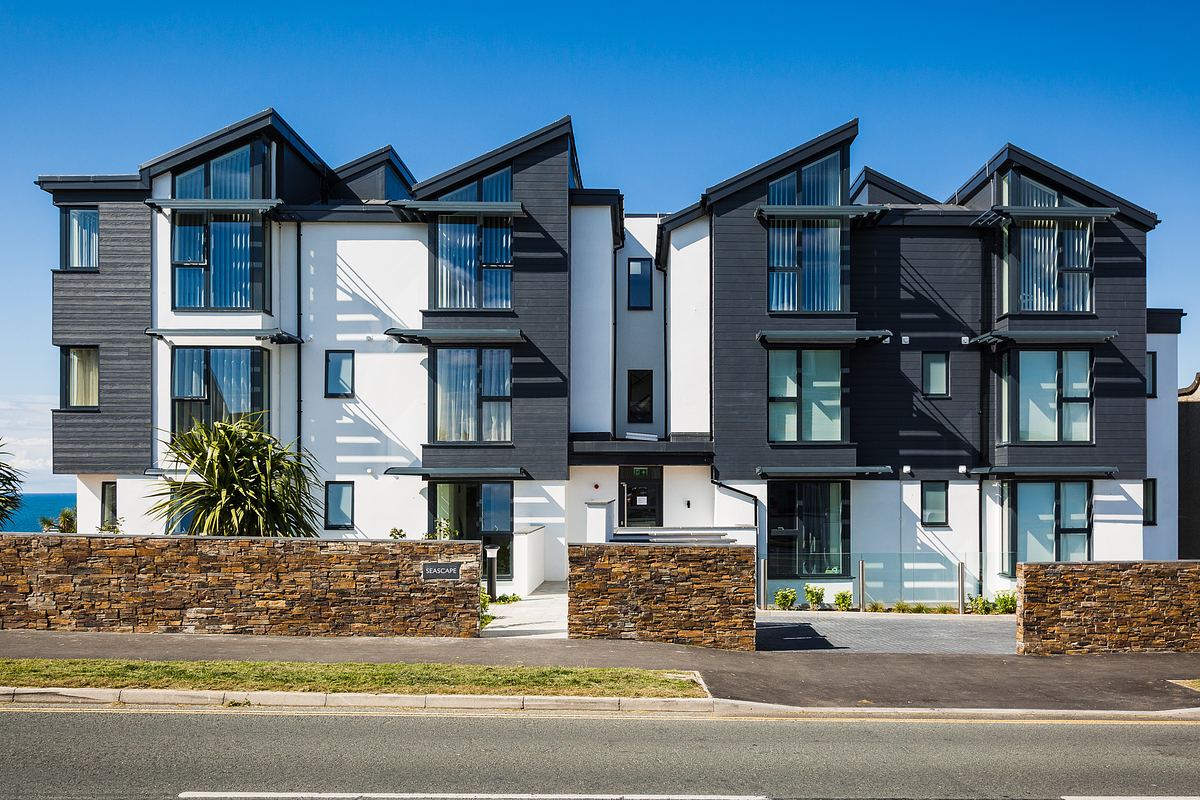Modern apartment building with angular roofs, vertical windows, and contrasting dark and light exterior. Stone wall and driveway in front under a clear blue sky.