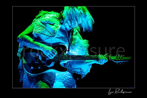 Horizontal color image of John 5 performing live on stage under blue and green lighting with a black electric guitar, white stage outfit, and tousled hair