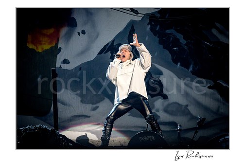 Bruce Dickinson of Iron Maiden performing live on stage in a white outfit, singing into a microphone with one hand raised against a dramatic dark stage backdrop.