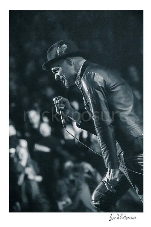 Vertical black and white image of Gord Downie of The Tragically Hip leaning forward with microphone in hand during a live performance, wearing a hat and leather jacket with the crowd softly blurred behind him