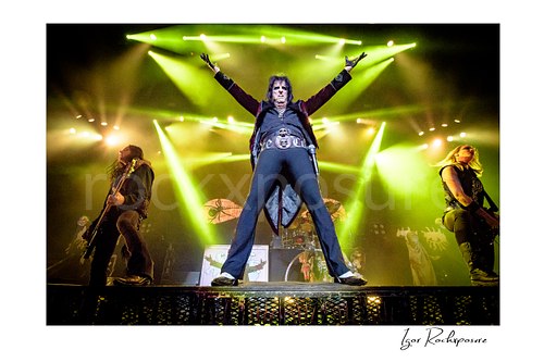 Horizontal color image of Alice Cooper, Chuck Garric and Nita Strauss of the Alice Cooper Band performing live on stage with raised arms, bright green lighting, and a dramatic low-angle perspective