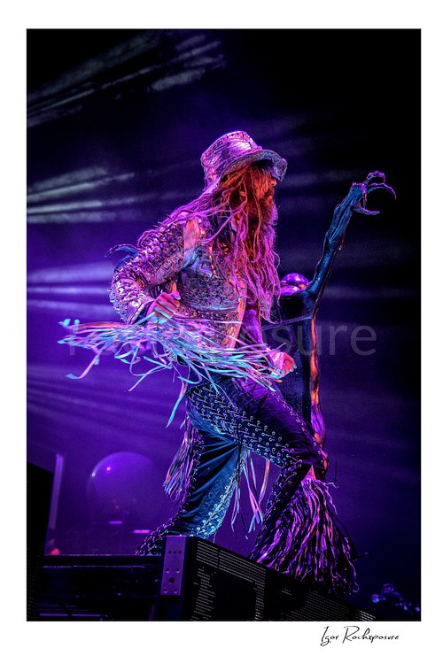 Vertical color image of Rob Zombie performing live on stage under purple lighting with a glittering hat, fringed jacket, dreadlocks, and a dark sculptural microphone stand