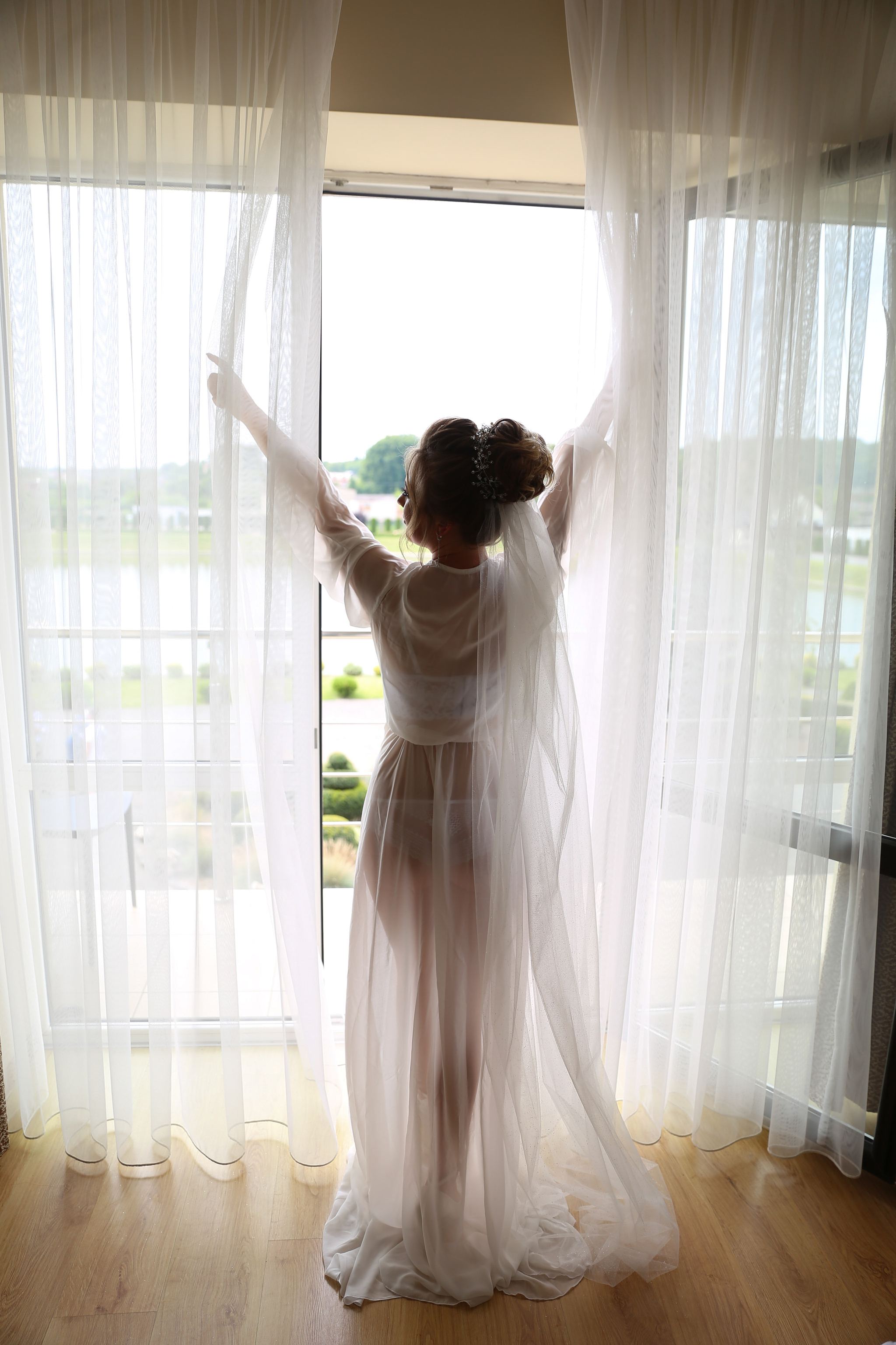 Bride opening sheer curtains on the morning of her wedding
