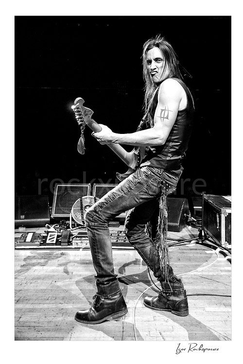 Vertical black and white image of Nuno Bettencourt performing live on stage with an electric guitar in a dynamic full-body pose
