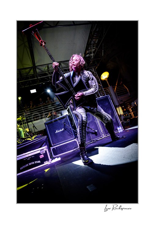 Vertical color image of Stig Pedersen of D-A-D performing live on stage in a striped outfit, holding his bass upright in a dramatic low-angle concert shot