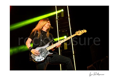 James LoMenzo of Megadeth smiles while playing bass with one knee raised, his long blond hair lit warmly against diagonal green stage beams.