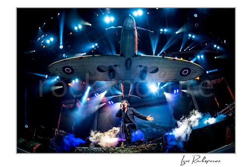Horizontal color image of Bruce Dickinson of Iron Maiden performing live on stage beneath the band’s large airplane stage prop with blue lights and smoke effects