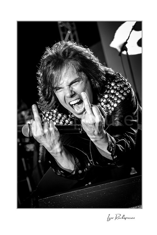 Vertical black and white image of Joey Tempest of Europe performing live on stage with a handheld microphone, studded jacket shoulders, open mouth scream, and both middle fingers raised