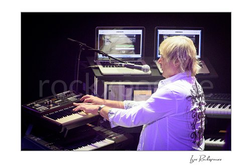 Horizontal color image of Geoff Downes of Yes performing at the keyboard rig with multiple synthesizers and screens during a live performance