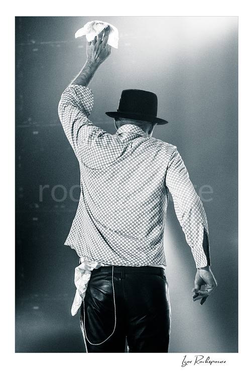 Vertical black and white image of Gord Downie of The Tragically Hip seen from behind on stage, raising a white cloth in a final farewell gesture under soft concert light