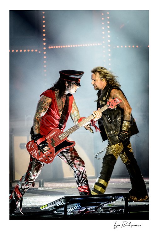 Vertical color image of Nikki Sixx and Vince Neil performing live on stage during a Mötley Crüe concert with bass guitar and stage smoke