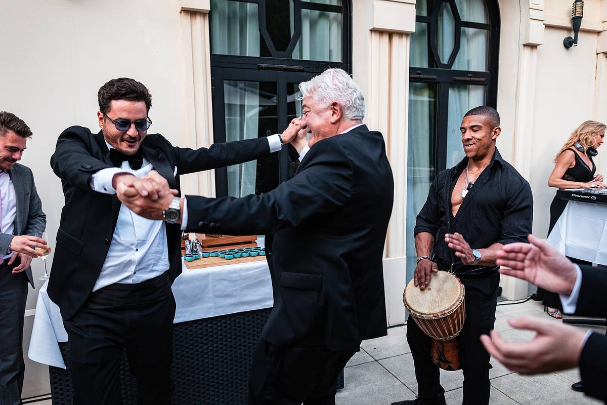 Le mari&eacute; et son p&egrave;re en tenue de soir&eacute;e partagent un moment de danse enthousiaste, entour&eacute;s de convives anim&eacute;s, dans un cadre ext&eacute;rieur lumineux, immortalis&eacute;s par Sebastien CLAVEL Photographe mariage Lyon