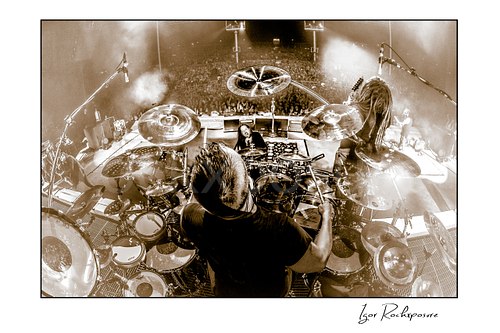 Horizontal black and white image of Korn drummer Ray Luzier seen from behind at the drums with the stage and crowd visible beyond him