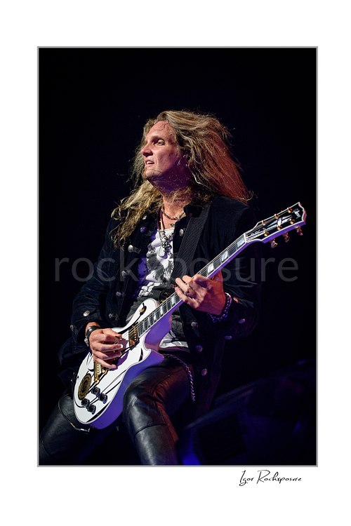 Vertical color image of Joel Hoekstra of Whitesnake performing live on stage, playing electric guitar under dramatic concert lighting