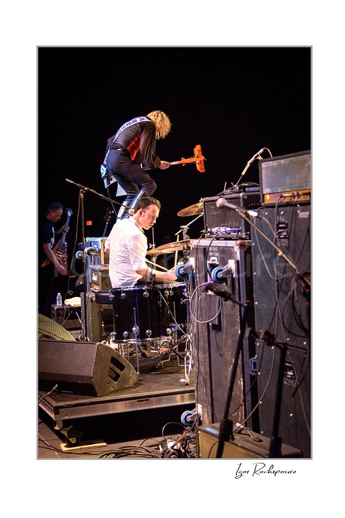 Vertical color image of Stig Pedersen leaping above drummer Laust Sonne during a D-A-D live performance, with drum kit, stage gear, and speaker stacks visible