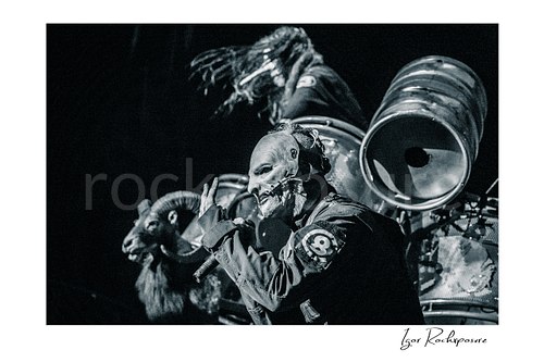 Horizontal black and white image of Corey Taylor of Slipknot wearing a mask and singing into a handheld microphone with industrial drums and a ram prop behind him