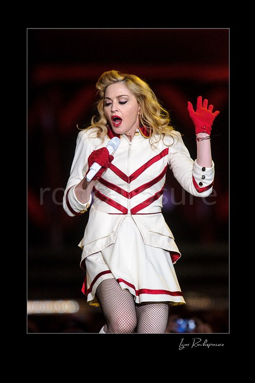 Vertical color image of Madonna singing into a microphone in a white and red stage outfit with one red-gloved hand raised