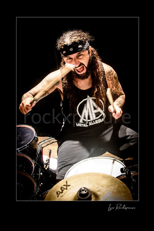 Vertical color image of Mike Portnoy at the drum kit with drumsticks raised and an intense expression under dramatic stage lighting