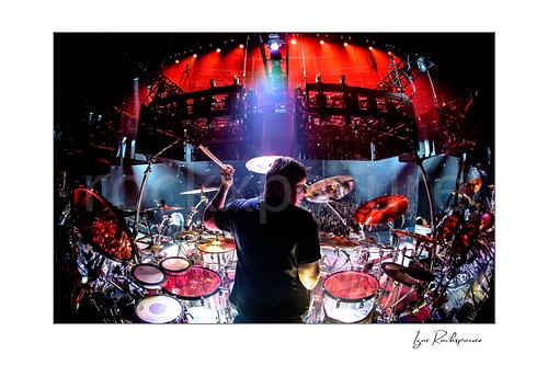 Horizontal color image of Korn drummer Ray Luzier seen from behind at the drums with one drumstick raised above the stage and crowd under vivid red lighting