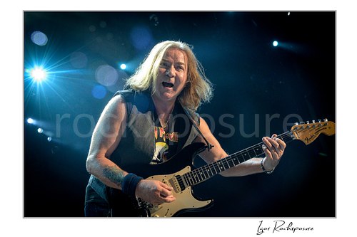 Horizontal color image of Dave Murray of Iron Maiden performing live on stage, playing electric guitar under blue concert lighting