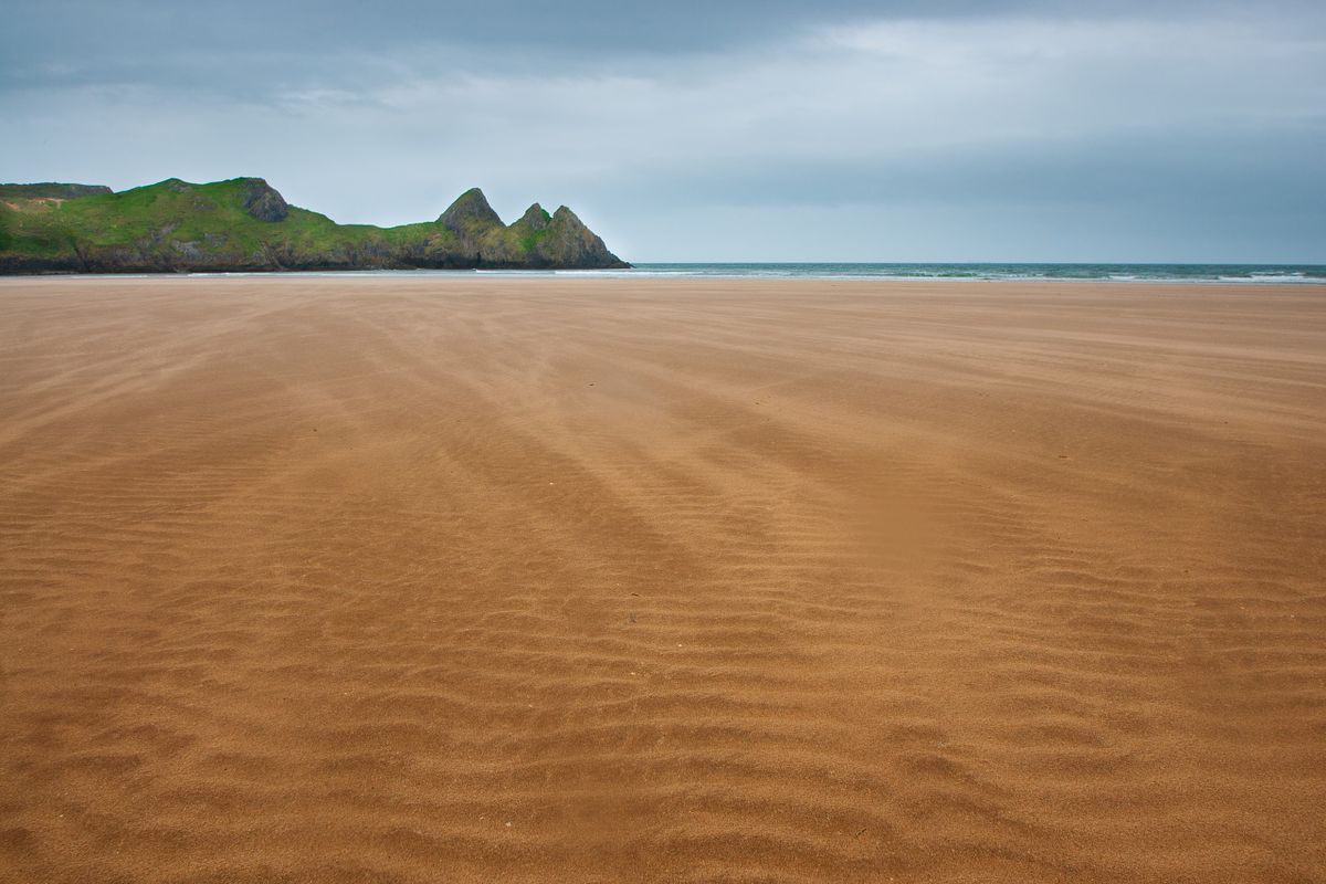 The wide sandy beach at Three Cliffs Bay on the Gower Peninsula, with the cliffs visible in the distance.