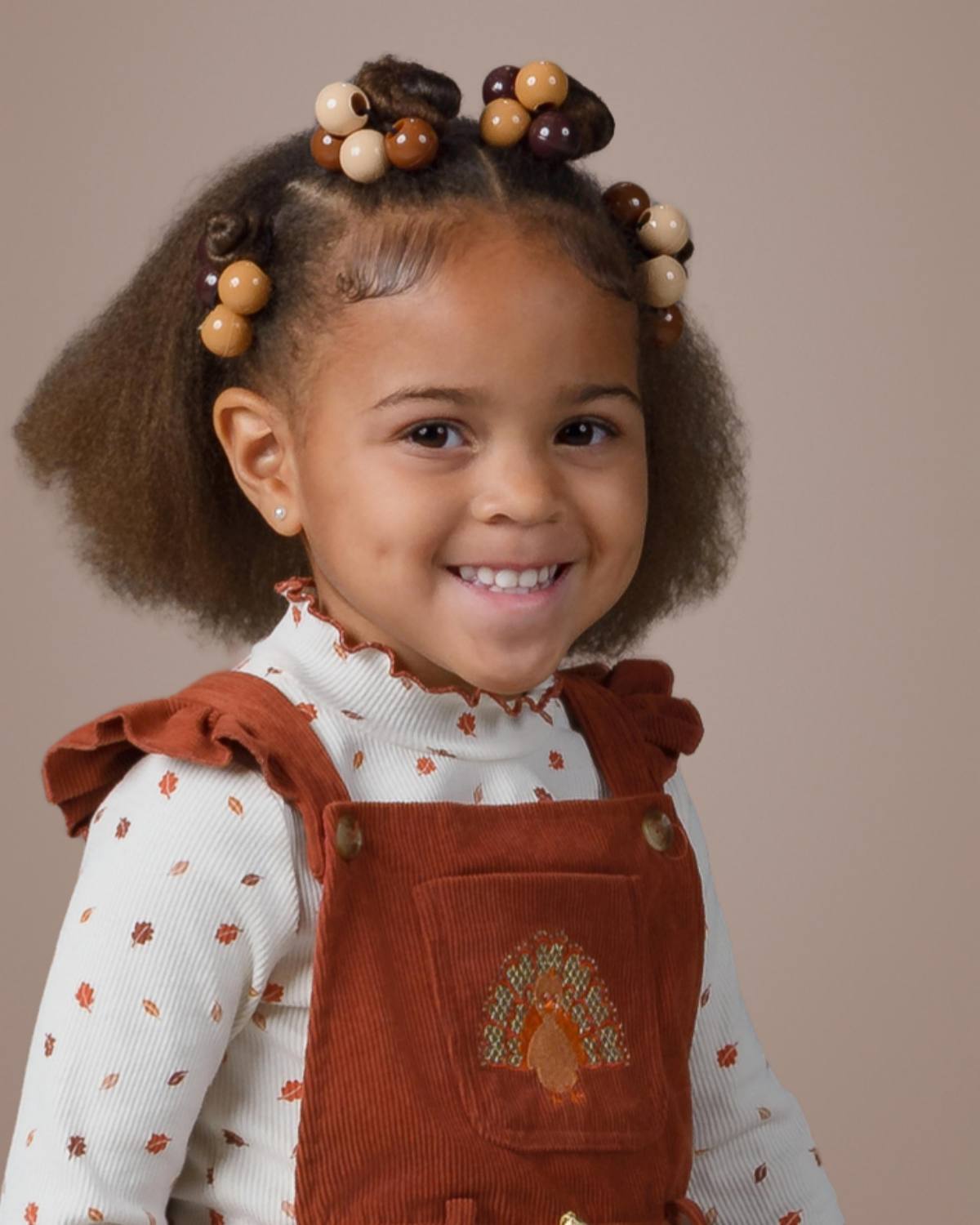 Preschool girl with shoulder-length hair styled in braids with round beads smiles at the camera, wearing rust-colored corduroy overalls over a cream patterned top, posed against a soft beige studio background for a class photo. | ProClicks Photography