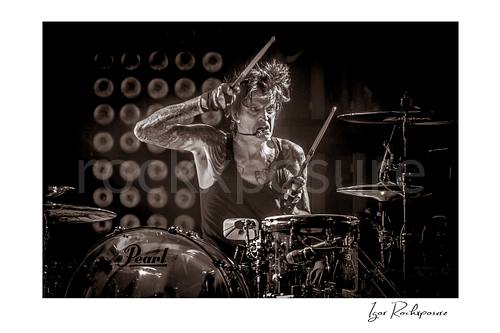 Horizontal black and white image of Tommy Lee performing at the drum kit during a Mötley Crüe concert, holding raised drumsticks under dramatic stage light