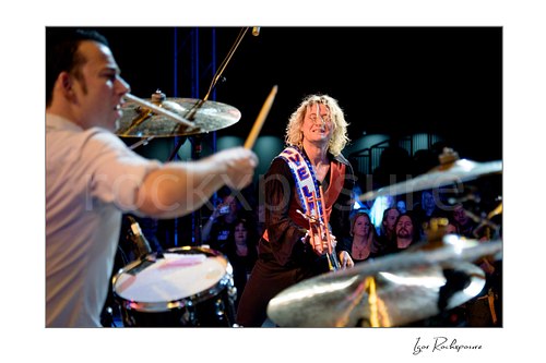Horizontal color image of Laust Sonne and Stig Pedersen performing live on stage, with Stig Pedersen smiling toward the crowd and drum cymbals framing the scene
