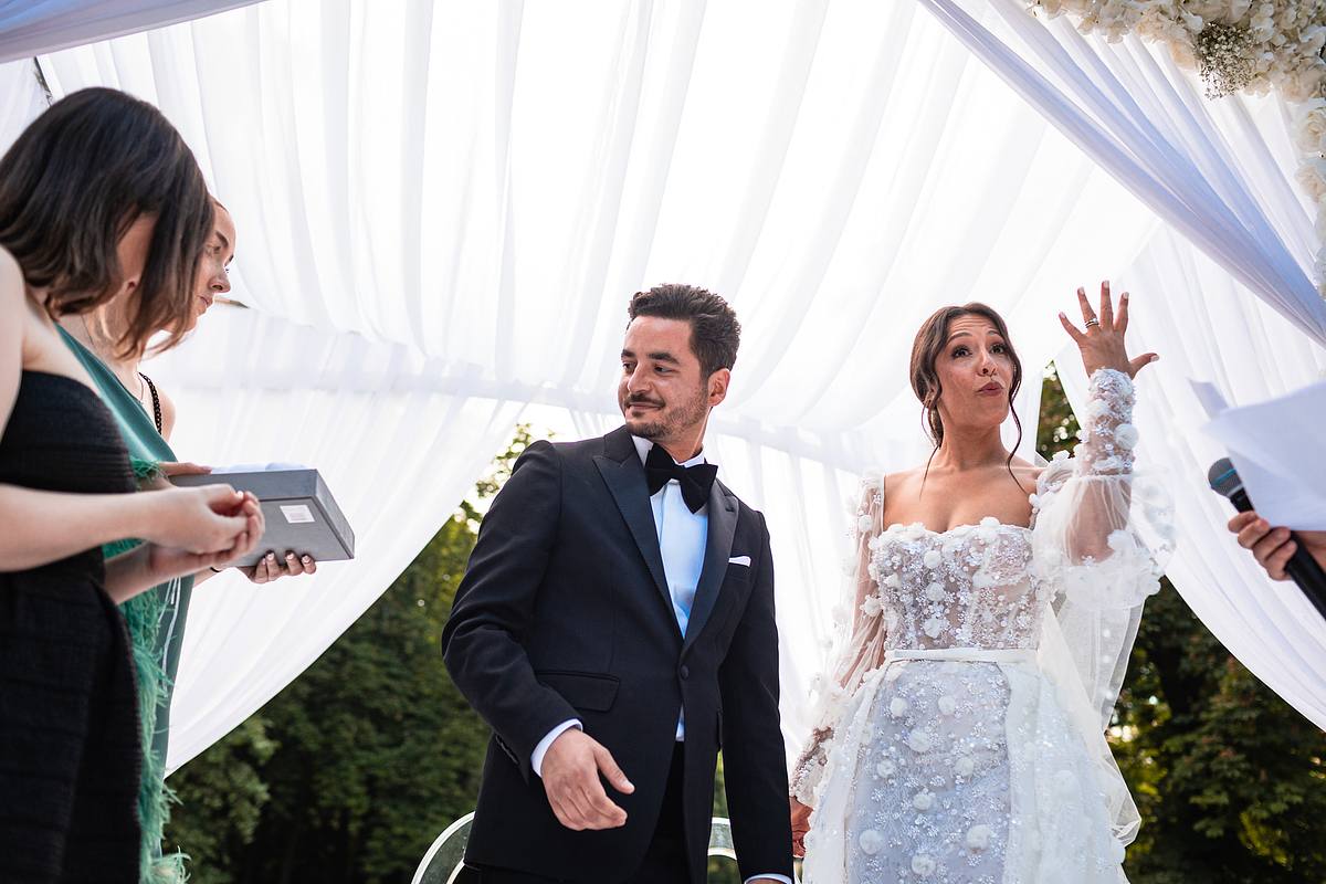 Sebastien CLAVEL Photographe mariage Lyon Un couple &eacute;l&eacute;gant debout sous une arche de mariage luxuriante, le mari&eacute; en costume noir et la mari&eacute;e en robe blanche d&eacute;taill&eacute;e, avec des invit&eacute;s applaudissant en arri&egrave;re-plan