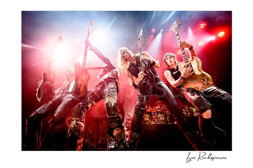 Horizontal color image of Alice Cooper, Chuck Garric, Nita Strauss, Tommy Henriksen and Ryan Roxie of the Alice Cooper Band performing live on stage with striped pants, guitars, and bright concert lighting