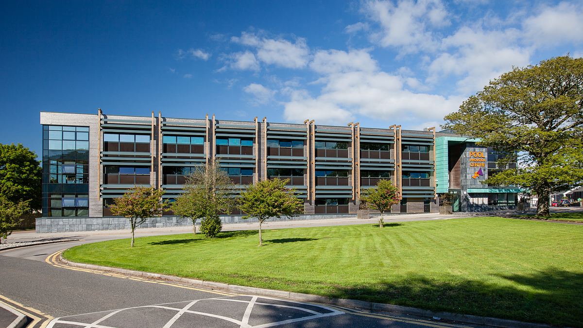 The front facade if a modern three-story office building with large windows, surrounded by green lawns and trees under a blue sky, perfect for an architectural photographer in Cornwall to capture its striking design.