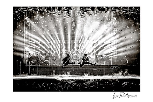Horizontal black and white image of Trans Siberian Orchestra performing on stage with two jumping guitarists, dramatic light beams, and a packed concert crowd