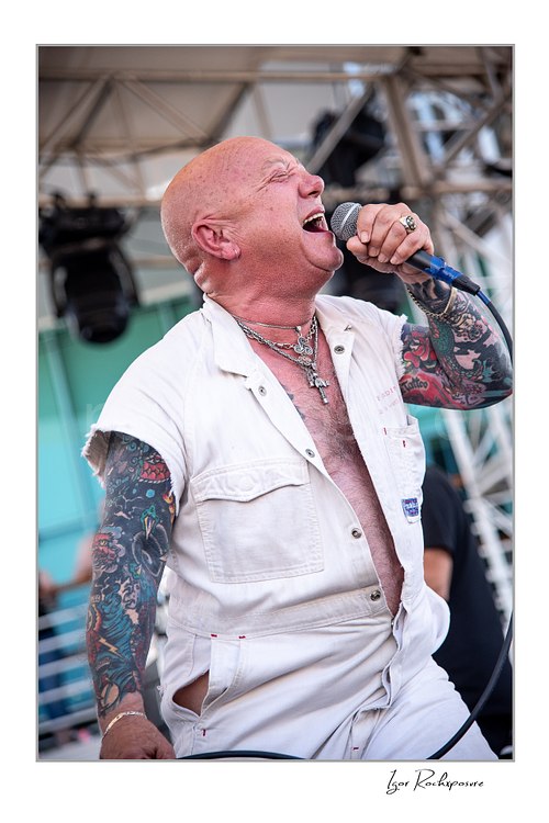 Vertical color image of Angry Anderson of Rose Tattoo singing into a microphone onstage in a sleeveless white outfit