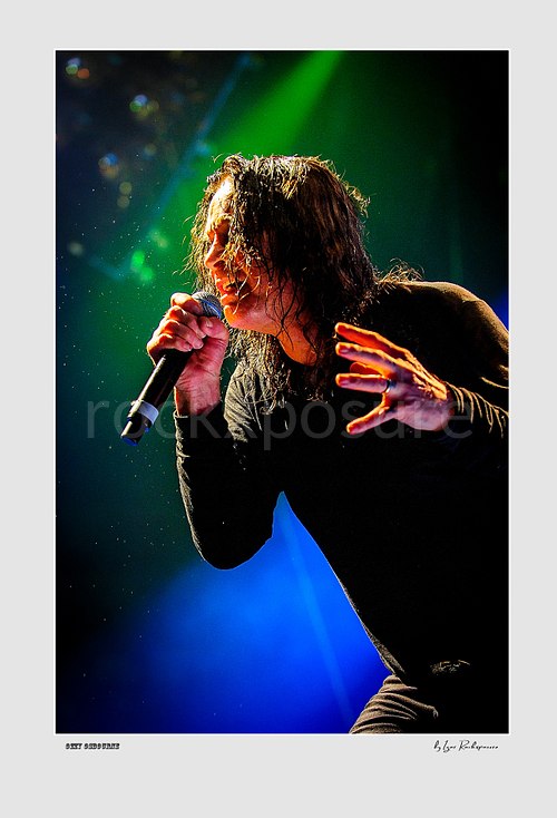 Ozzy Osbourne of Black Sabbath singing into a microphone with expressive hand gesture under dramatic green and blue stage lighting