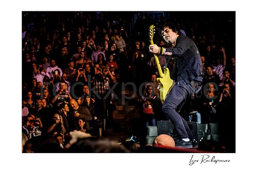 Horizontal color image of Billie Joe Armstrong of Green Day playing guitar on a stage riser with a large crowd behind him
