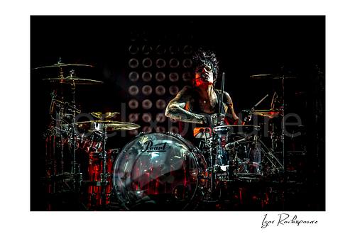Horizontal color image of Tommy Lee performing at the drum kit during a Mötley Crüe concert, framed by cymbals with dramatic red stage light
