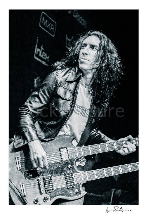 Vertical black and white image of Brent Woods playing double-neck guitar on stage in a tight performance shot