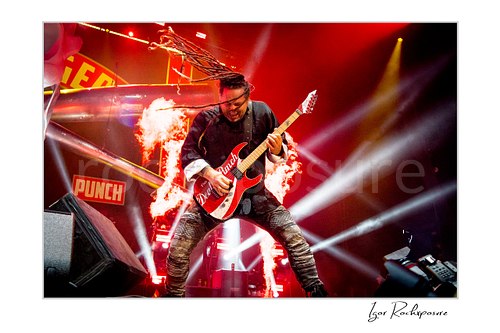 Horizontal color image of Jason Hook of Five Finger Death Punch playing guitar onstage with flying dreadlocks, flames, and bright concert beams