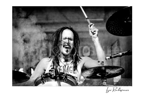 Horizontal black and white image of Ginger Fish performing live on stage at the drums with raised drumstick, flying hair, and drifting stage smoke