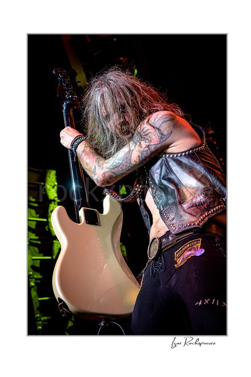 Vertical color image of Jan LeGrow of Beasto Blanco gripping a white bass guitar in a powerful live performance pose, with long hair covering his face and tattooed arms under warm stage light