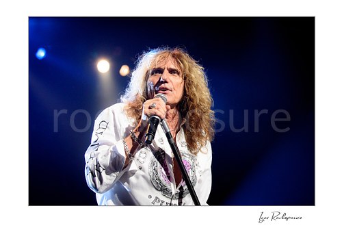 Horizontal color close-up image of David Coverdale of Whitesnake performing live on stage, singing into a microphone while holding a mic stand under blue concert lighting