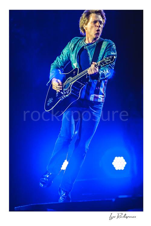 Vertical color image of Jon Bon Jovi performing live with a black acoustic guitar under vivid blue stage lighting