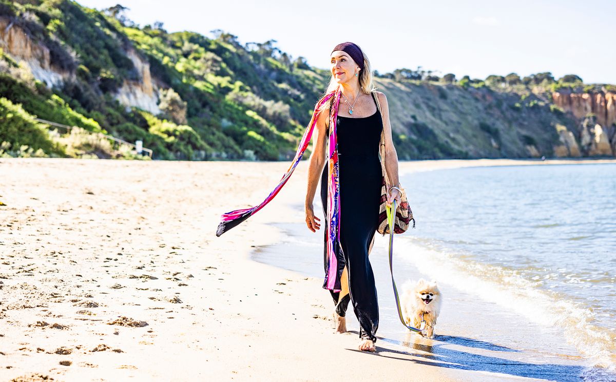A woman walks her little dog on Sandringham Beach. She has a long, colourful scarf on.