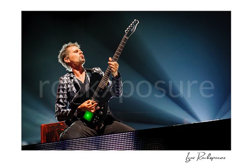 Horizontal color image of Matt Bellamy of Muse playing guitar in a patterned stage jacket with radiant blue stage beams behind him