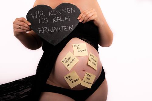 Frontale Nahaufnahme einer Schwangeren in schwarzer Unterwäsche mit fliegender Schleppe; sie hält ein Schieferherz mit der Aufschrift ‚Wir können es kaum erwarten‘ vor den Bauch, der mit beschrifteten Post-its beklebt ist.