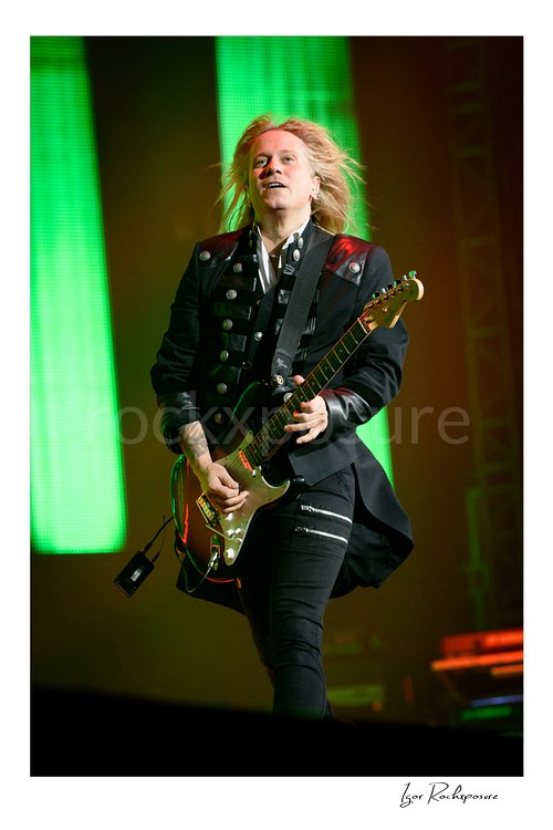 Vertical color image of Chris Caffery of Trans Siberian Orchestra playing electric guitar with windblown hair against green stage lighting
